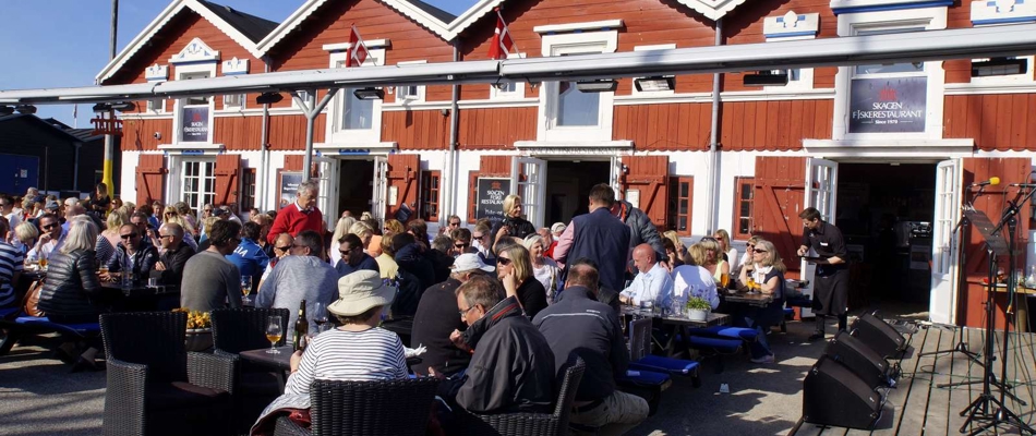 Mange mennesker sidder udendørs foran de røde havnebygninger i Skagen og nyder mad og drikke i solskin